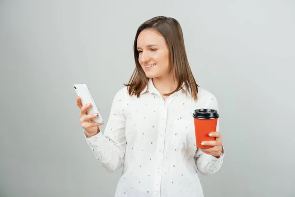 年轻的微笑的女人看着电话 手里拿着一杯咖啡 斯托迪奥在灰蒙蒙的背景下拍摄 — 图库照片