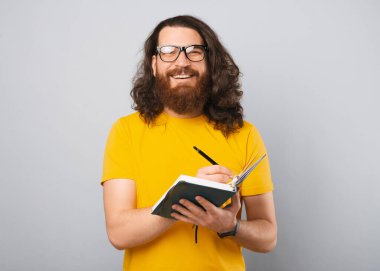 Uzun saçlı neşeli genç adamın fotoğrafı ajandasında notlar alıyor ve gülümsüyor..