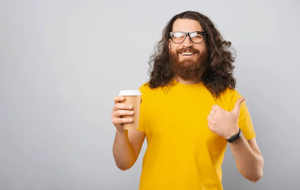 一个留着长发大胡子的快乐年轻人的画像 他伸出大拇指 拿着纸杯咖啡 — 图库照片