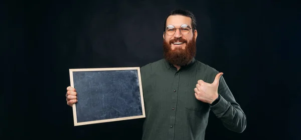 Usmívající se vousatý muž drží černou tabuli, zatímco ukazuje palec nahoru. — Stock fotografie