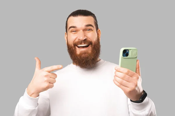 Extático homem barbudo surpreso está segurando o telefone enquanto aponta para ele. — Fotografia de Stock
