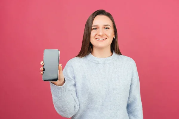 Jovem mulher sorrindo está segurando um telefone e mostrando tela para a câmera. — Fotografia de Stock