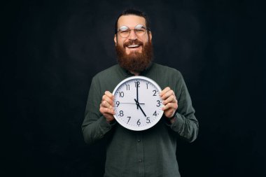 Mest olmuş sakallı adam gülümserken yuvarlak beyaz bir duvar saati tutuyor..