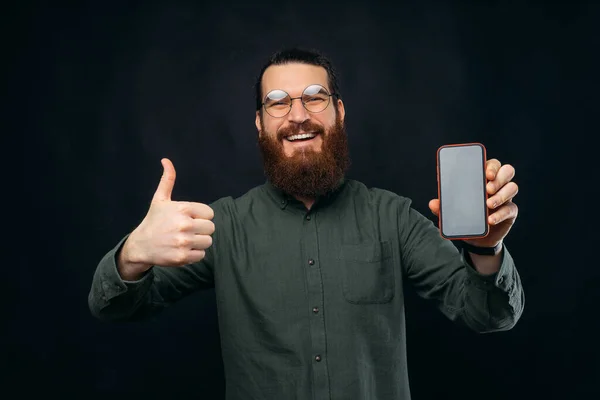 Hombre barbudo joven está mostrando a la cámara en blanco teléfono de la pantalla a un pulgar hacia arriba. — Foto de Stock