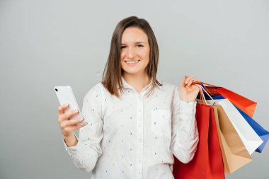 Cheerul genç bayan kağıt alışveriş torbaları tutuyor ve akıllı telefonuyla internetten bir şeyler alıyor.