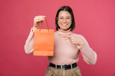 Mutlu genç bayan elinde turuncu bir alışveriş çantası tutuyor ve onu işaret ediyor. Fotokopi alanı ile pembe arkaplan üzerinde çekim.