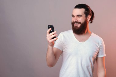 Handsome bearded man is looking on a phone while smiling wide. Studio shot over grey background with red light.