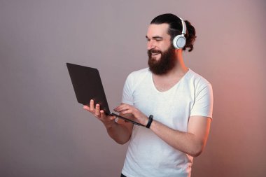 Kulaklık takan zeki genç adam gri arka planda elinde dizüstü bilgisayarla gülümsüyor..