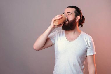 Susamış genç sakallı adam gözleri kapalı bir fincan kahve içiyor. Stüdyo görüntüsü gri arkaplan üzerinde kırmızı ışık.