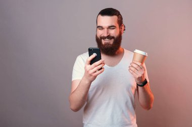 Smiling man is surfing the internet on phone while holding a coffee cup. Studio whot with red light.