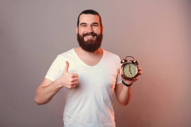 Mutlu sakallı adam elinde çalar saat tutarken jest yapıyor. Stüdyo görüntüsü gri arkaplan üzerinde kırmızı ışıkla.