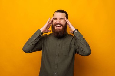 Genç sakallı adam baş ağrısı yüzünden el ele tutuşuyor. Stüdyo çekimi sarı arkaplanda.