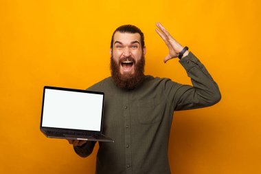 Ecstatic bearded man holding a laptop with blank screen is mind blown. Studio shot over yellow background.
