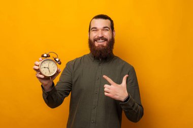 Mutlu sakallı adam elinde tuttuğu çalar saati işaret ediyor. Stüdyo çekimi sarı arka planda..