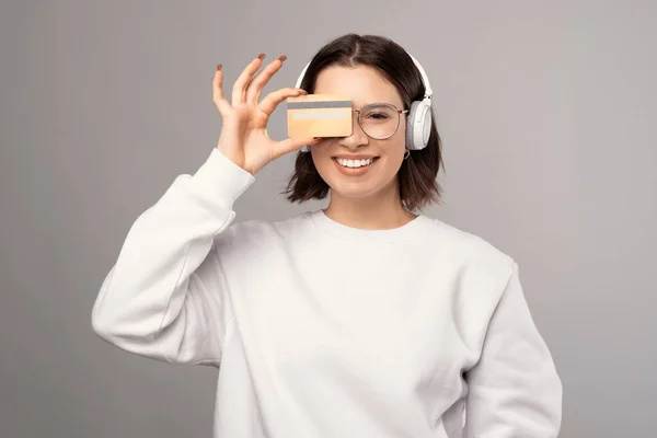Smiling woman is holding a credit card and hiding behind while wearing headphones. — 스톡 사진