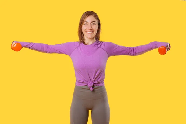 Young smiling woman is exercising arms with dumbbells over yellow background. — Fotografia de Stock