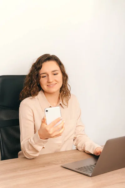 Vertical photo of charming young woman sitting on desk in office and using smartphone. — Foto de Stock