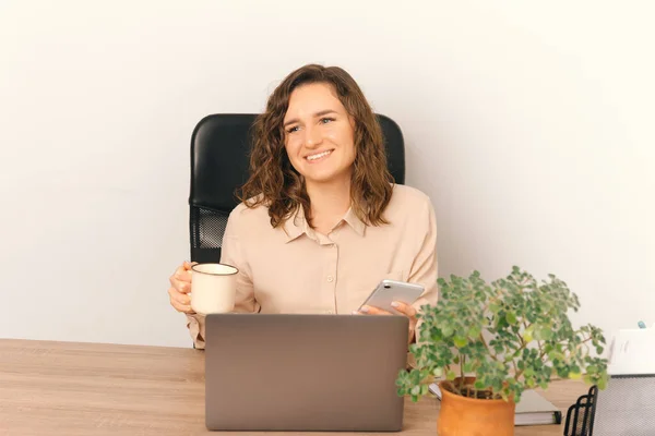Photo of joyful young office woman in casual sitting at desk and holding cup of coffee and smartphone. — Stockfoto