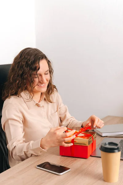 Vertical portrait of smiling young woman sitting at office table and opening red gift box. — Stockfoto