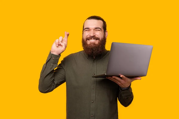 El hombre barbudo positivo sostiene un portátil abierto mientras cruza los dedos. — Foto de Stock