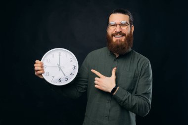 Yuvarlak gözlüklü genç sakallı adamın fotoğrafı koyu arkaplanda beyaz saati gösteriyor..