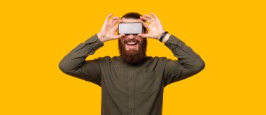 Gülümseyen sakallı adam, sarı bir arka planın yanında duran bir telefonu gözünün üstünde tutan adamdır.