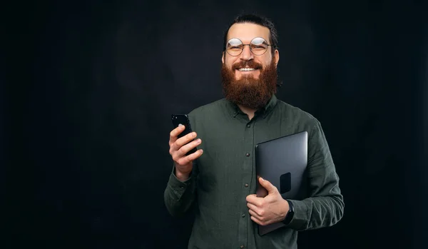 A happy bearded man is holding a laptop and his phone and is smiling is looking at the camera
