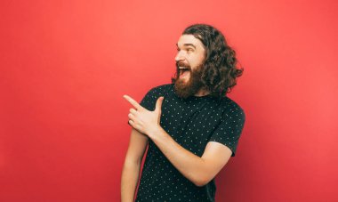 A happily young bearded man is pointing at some free space while looking at it