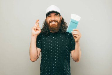 An excited yung man is holding his passport and some tickets in it while he is standing with his fingers crossed