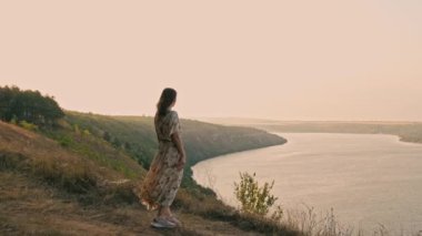 Beautiful footage of woman standing on windy hill near big river at sunset and enjoying nice weather.