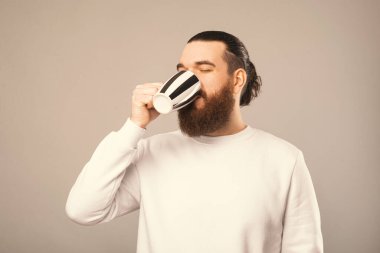 Bearded hipster wearing white sweatshirt enjoys his warm beverage from a mug with white and black stripes. Studio photo over grey background.