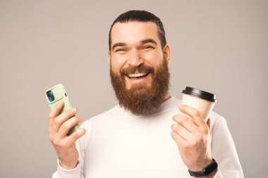 Man wide smiling at the camera is holding a take away coffee cup and a phone. Close up studio portrait over warm grey background.