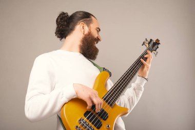 Ecstatic young bearded man wearing white sweatshirt is playing bass in a studio over grey background. Low angle shot.