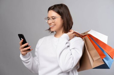 Cheerful young woman wearing glasses looks at the phone while holding shopping bags. Studio shot over grey backdrop.