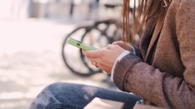 footage of casual woman sitting on bunch and writing text in smartphone in city.