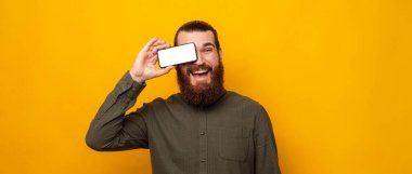 Playful bearded man covers one eye with phone screen and smiles at the camera. Studio portrait over yellow background.