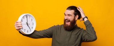 Terrified bearded man with hand on head is looking at the white round clock he is holding because time flys fast. Studio shot over yellow background.