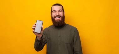Handsome bearded man shows phone screen to the camera over yellow background. Studio portrait shot.