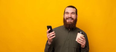 Cheerful bearded man smiles at the camera while holding a to go cup and a phone. Studio shot over yellow background.