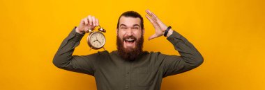 Bearded man is mad because time flys so fast while holding an alarm clock. Studio portrait over yellow backdrop.