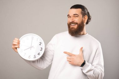 Playful bearded man is pointing at the white round clock he is holding while looking and smiling at the camera.