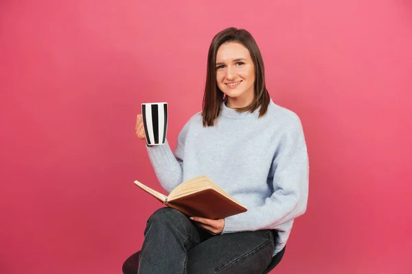 Brunette smiling at the camera woman is sitting on a chair with a book and mug. Studio shot over pink background.