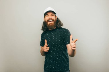 Hey sen, uzun saçlı, gri arka planda kameraya bakan ve gülümseyen genç sakallı adam..