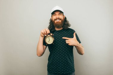 Mutlu genç hippi sakallı adam gri arka planda çalar saati işaret ediyor..