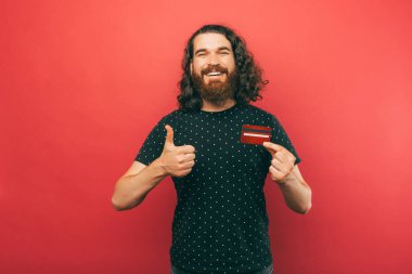 Tişörtlü mutlu sakallı adam başparmağını kaldırıyor ve elinde kırmızı banka kartı tutuyor.
