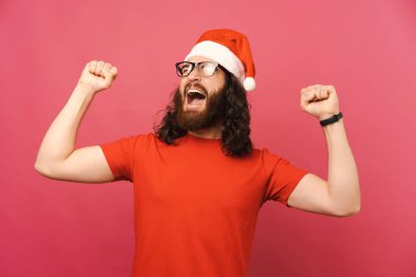 Noel Baba şapkası takan mutlu sakallı genç adamın fotoğrafı. Zaferi kollarını kaldırarak kutluyor..