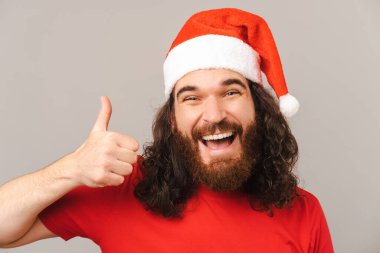 Noel şapkası takan sakallı bir adam baş parmağıyla onayını veriyor. Stüdyo çekimi gri arkaplan üzerinde.