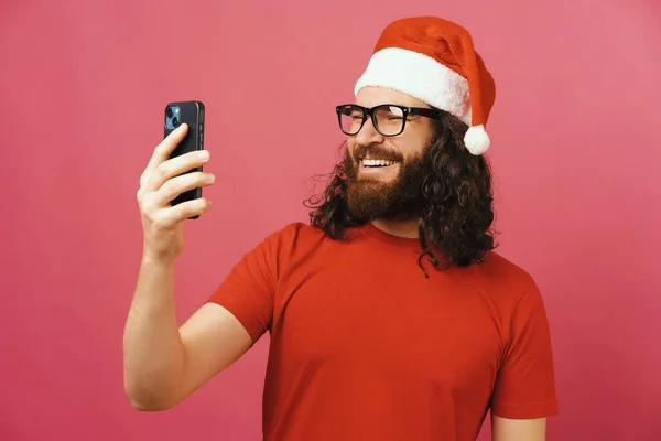 幸せな男はクリスマスの帽子をかぶっている間 自分の携帯電話で自撮りしています スタジオショットオーバーピンクの背景 — ストック写真
