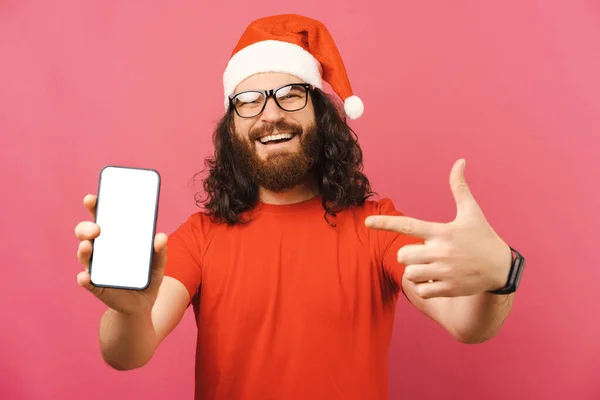 彼が持っている空の電話画面でクリスマスキャップポイントを身に着けているかわいい髭の男 スタジオショットオーバーピンクの背景 — ストック写真