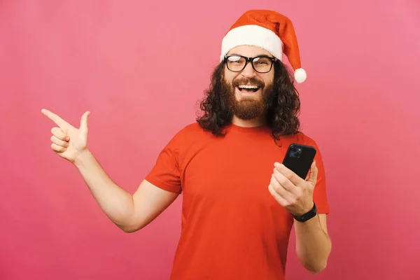 若いクリスマスの男は電話と広い笑顔を保持しながら脇にポイントします スタジオショットオーバーピンクの背景 — ストック写真